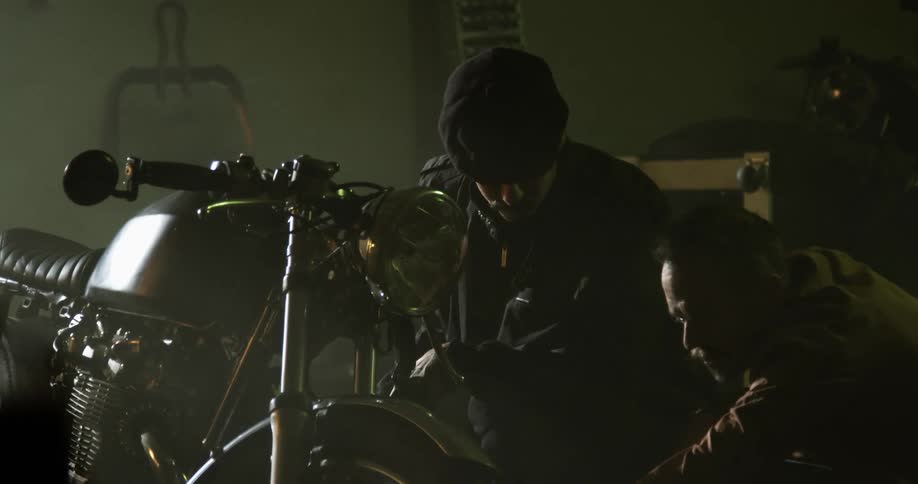 Two white men kneel by a motorcycle in a garage and talk