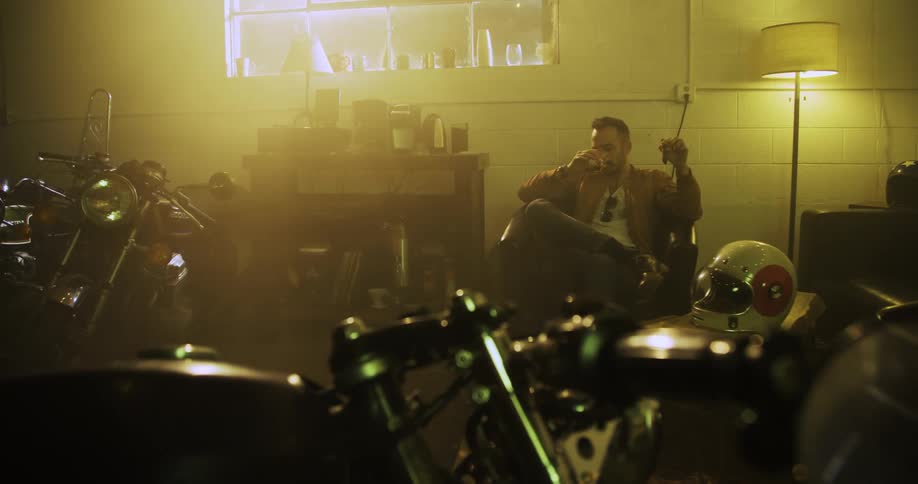 A man sits in a room with motorcycles and smokes and drinks