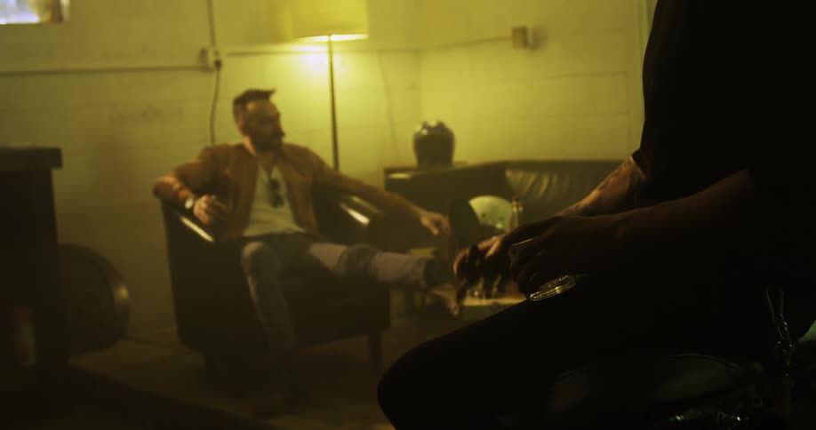 Two white men sit in a dingy living area smoking and drinking