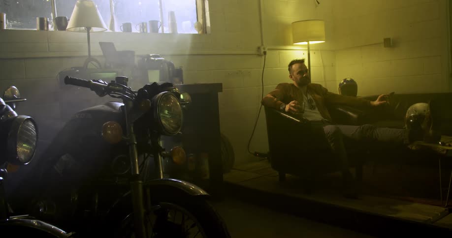 A man sits on a couch in a dim basement with a motorcycle inside