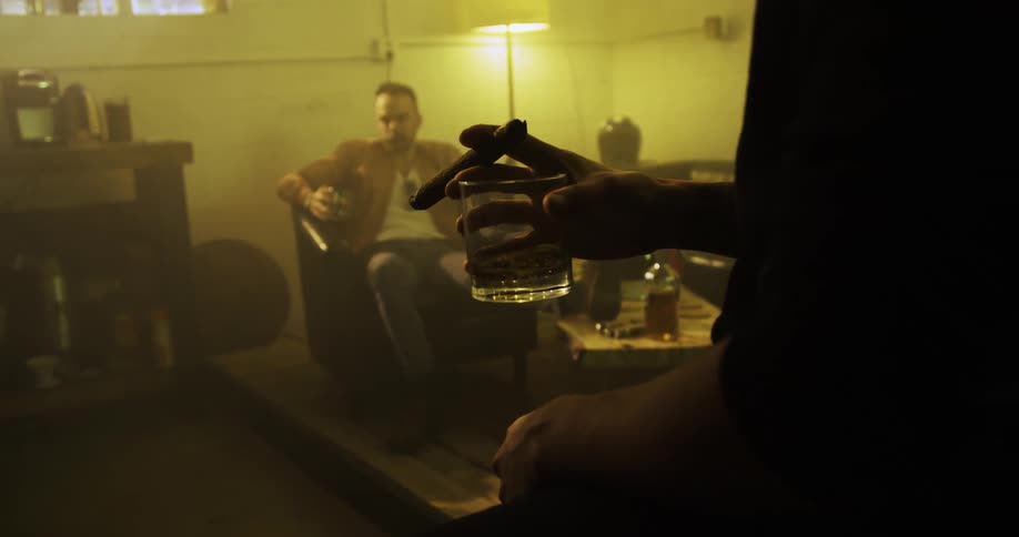 A man swishes liquor in a glass and holds a cigar while another man sits in the background drinking