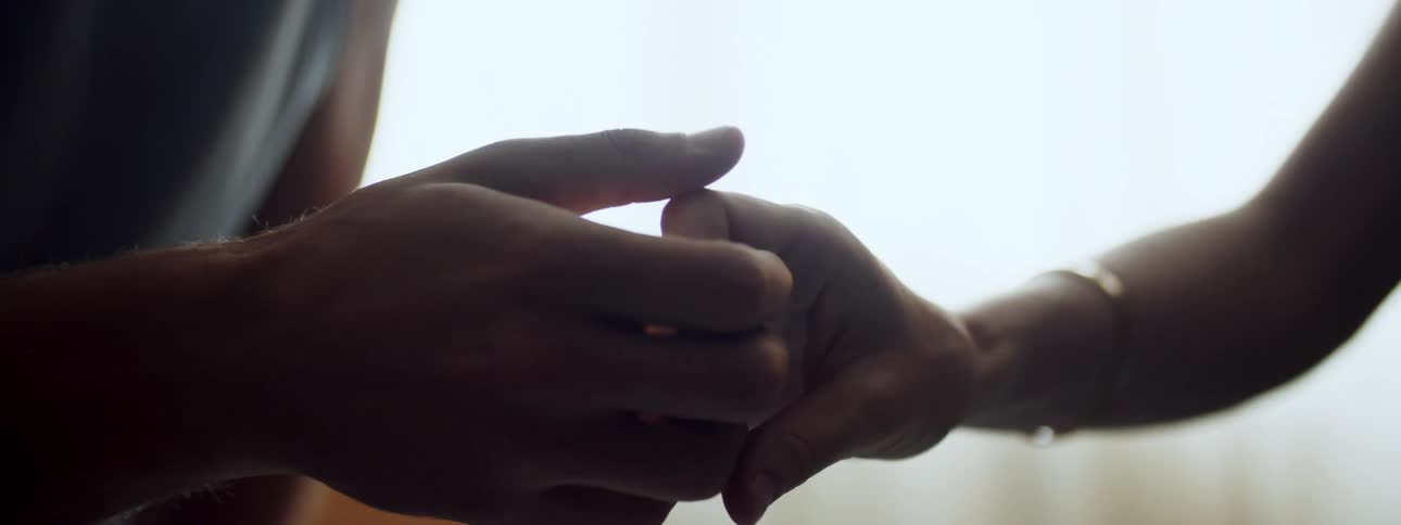 Woman with a gold bracelet sitting in a chair and man sitting on a bed hold each others hands
