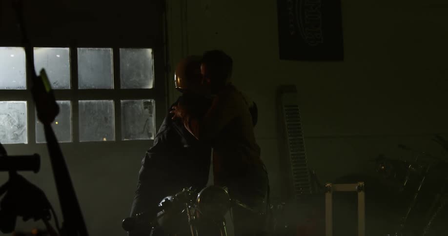 Two men greet each other inside a dimly lit motorcycle workshop