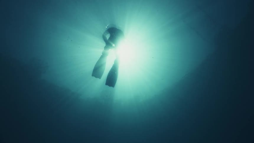 The silhouette of a diver wearing flippers floating near the surface where the sun is shining down