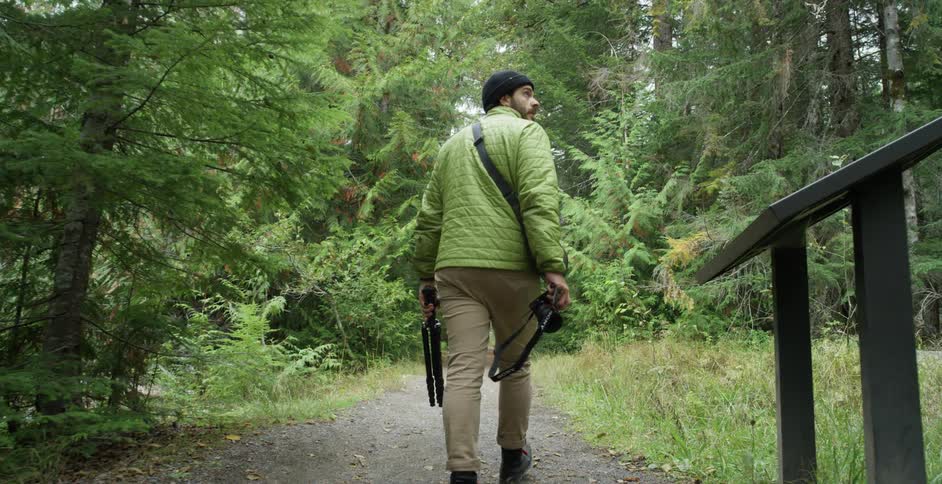 A white man totes a tripod and camera down a forest path