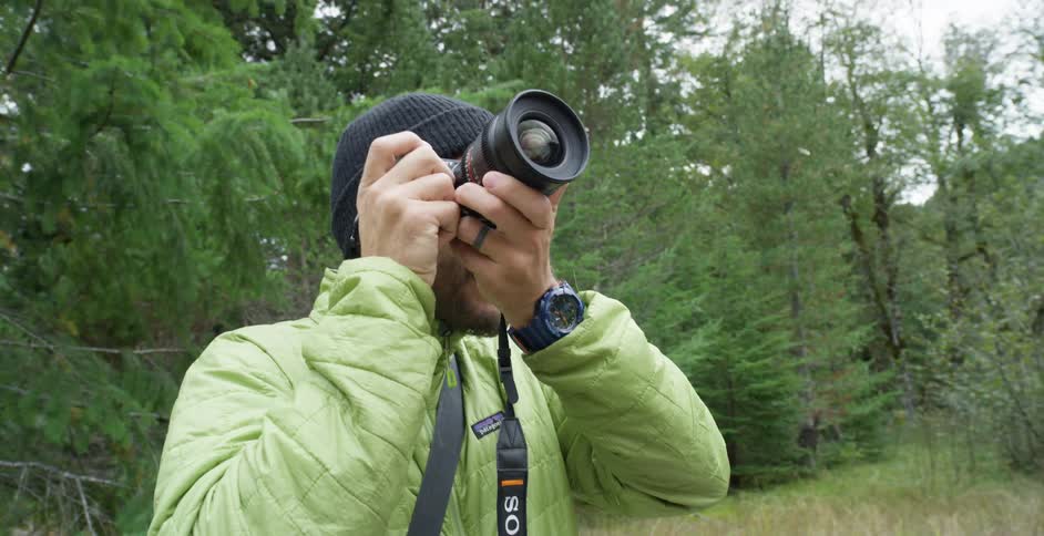 A photograph raises a camera to his face and adjusts the lens to take a photo in a woodland