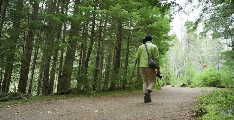 A white man with a camera walks down a path through a forest