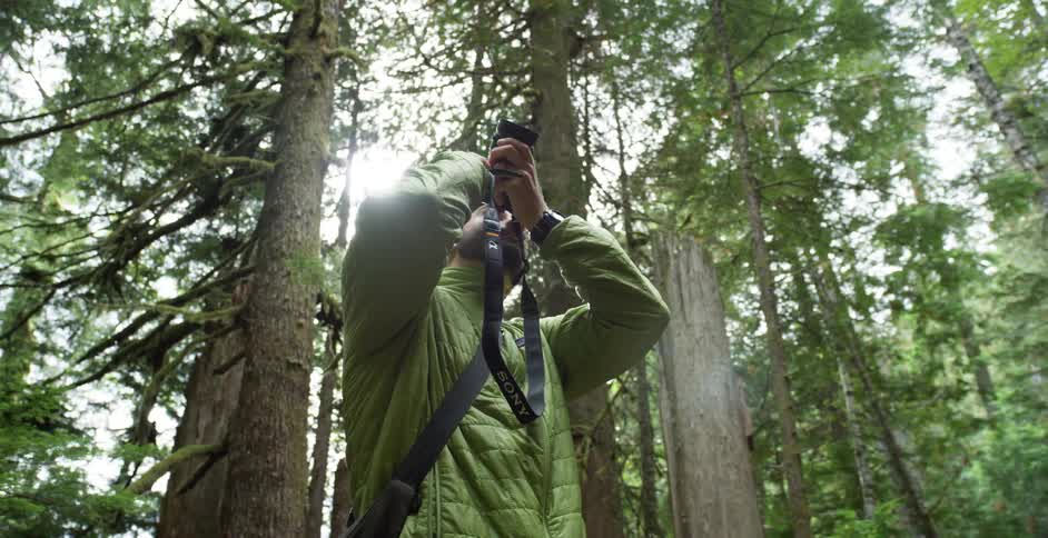 A white man in a green coat takes a picture in a forest with a camera