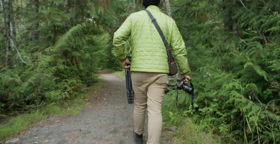 A white male with a camera and tripod walks on a forest path