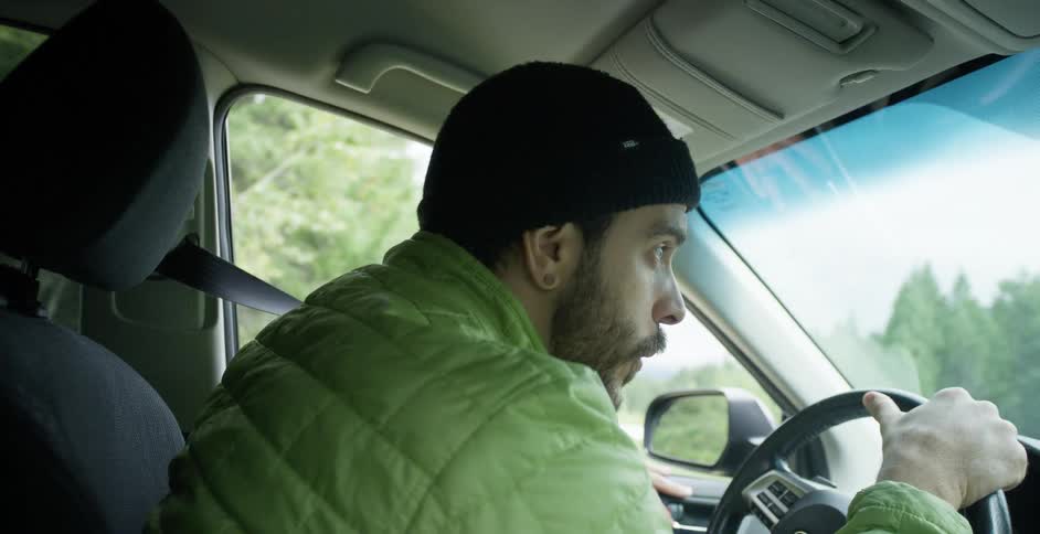 Man wearing coat and knit cap looks around while driving through forest