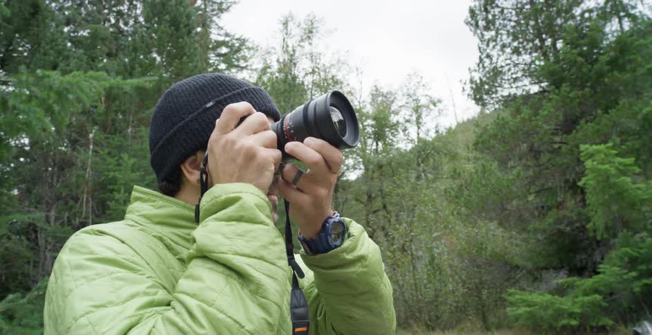 A man with a camera taking pictures in the woods with trees and the sky in the background