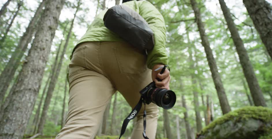 A white man carrying a tripod and camera walks through a forest