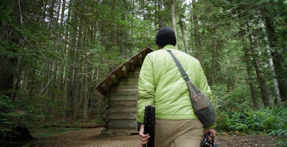 A man holds a camera and tripod and walks to a small cottage in the woods