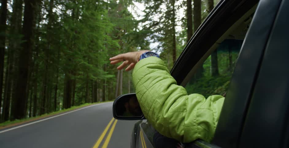 A man driving a car on a forest road holds his hand out the window playing in the wind