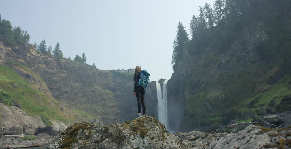 A white woman stands on a rock and looks at a waterfall in the distance