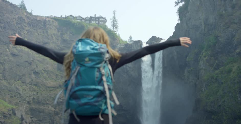 Girl with backpack stands in front of a tall waterfall in the mountains
