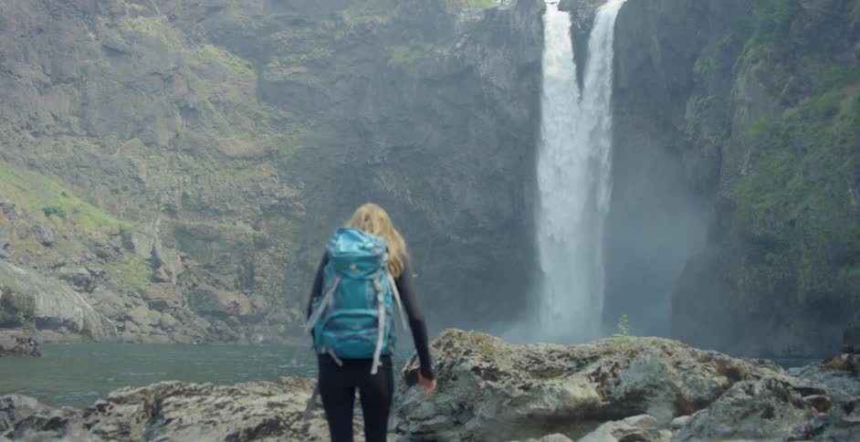 Young girl with backpack walks on rocks near a waterfall in the mountains