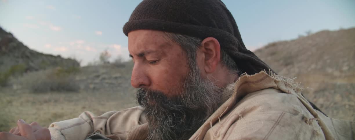 Bearded man sits in the ground with eyes closed murmuring to himself