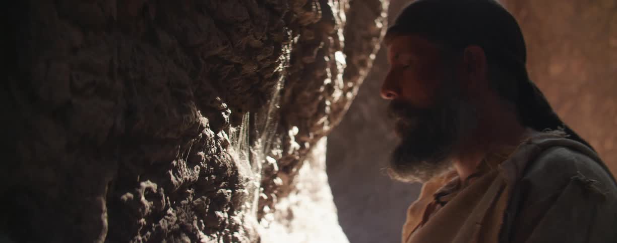 Man wearing robe and head covering prays next to shadowy rock wall partly lit by sunlight