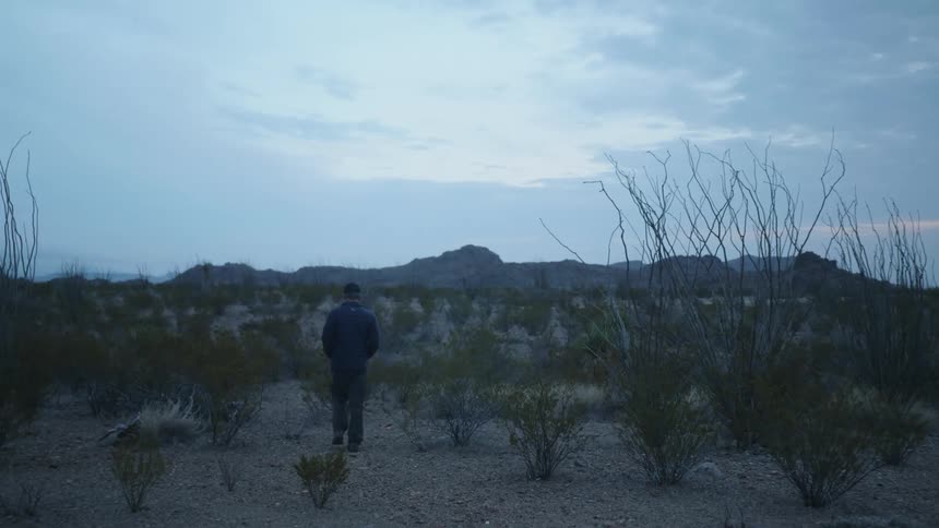 A person with a jacket walking in the desert with mountains the sky and clouds in the background