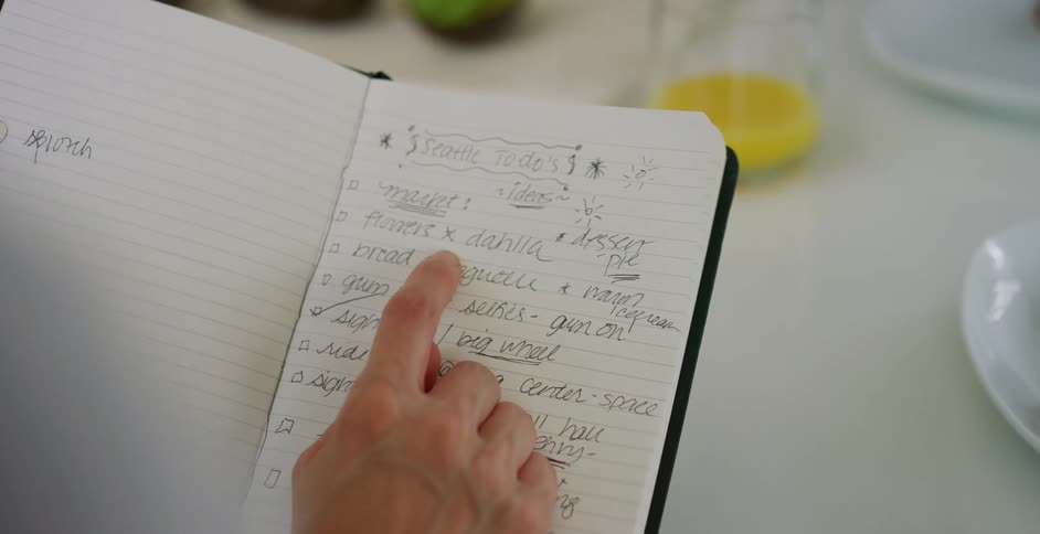 A person holds and points at a notebook with a to do list while another eats avocado toast