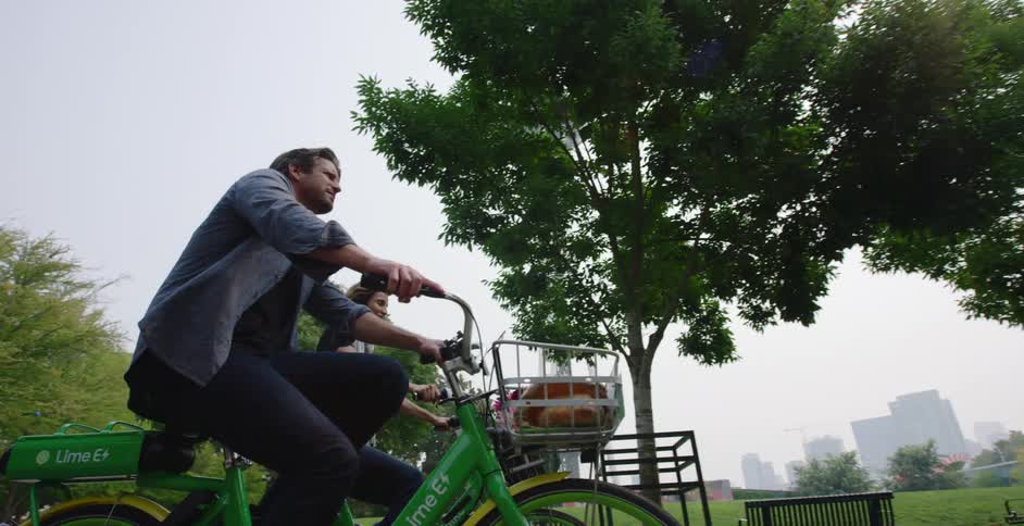 Couple rides bicycles through park with skyline in background