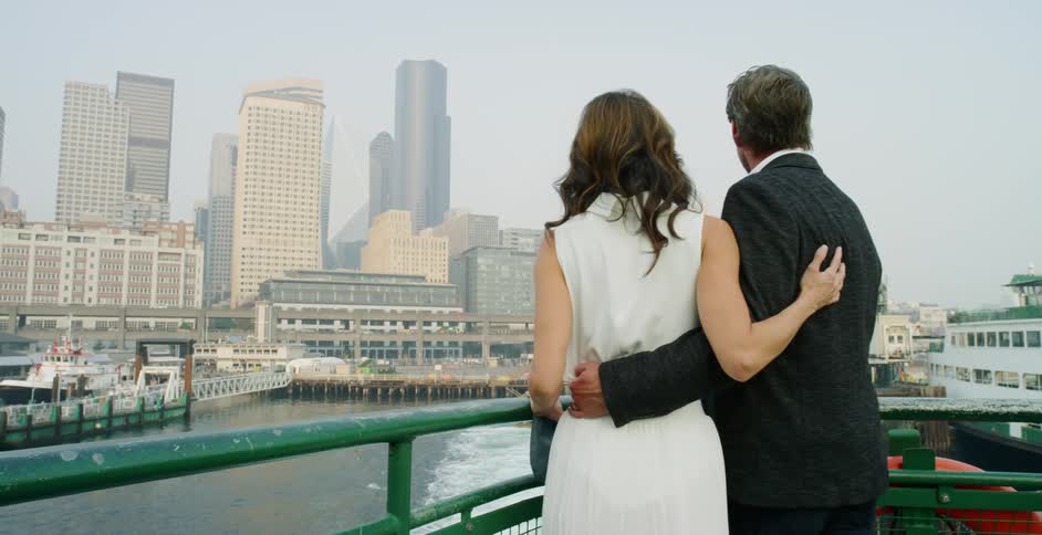 Couple stands at rear of ship overlooking mist covered city and wake