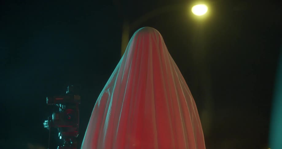 A figure covered in a shroud stands outdoors by a video camera