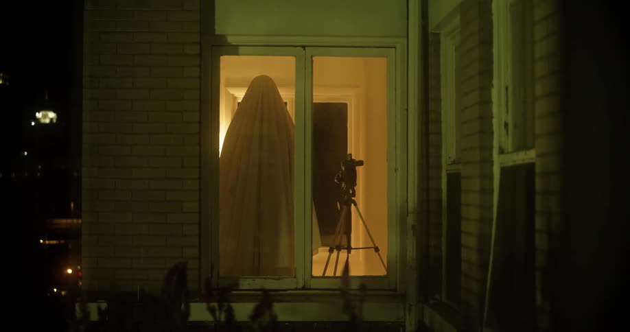 A person inside a home covered in a white sheet stands in front of a camera