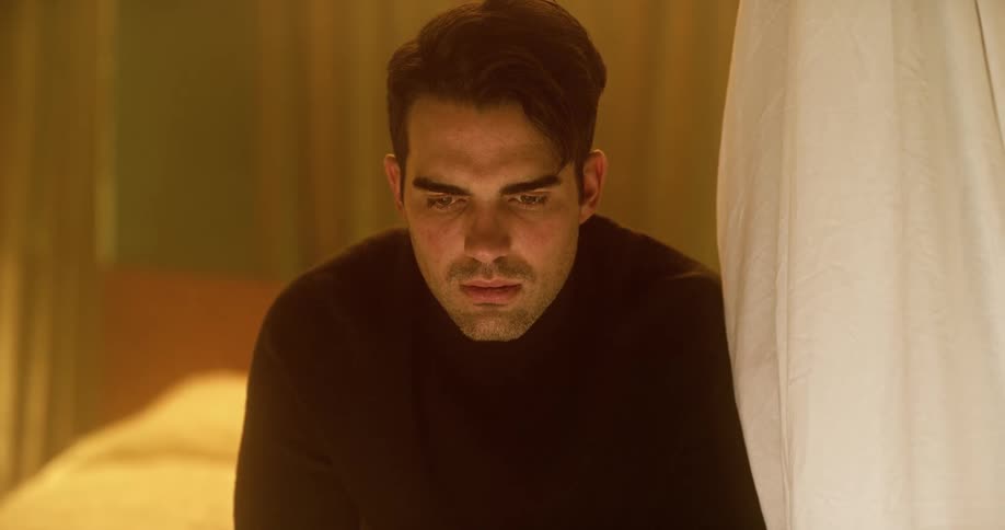 Man with a black sweater feeling depressed sits on a bed and stares blankly ahead in the bedroom