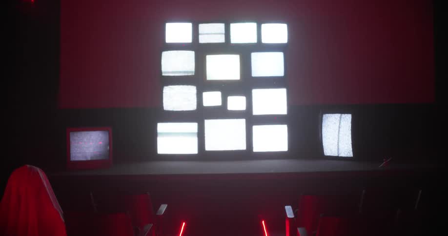 White sheet shrouded figures sit in dimly lit theater facing wall of televisions showing static