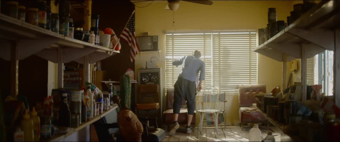 A man standing in a room with crowded shelves parts the blinds of a window to peak outside