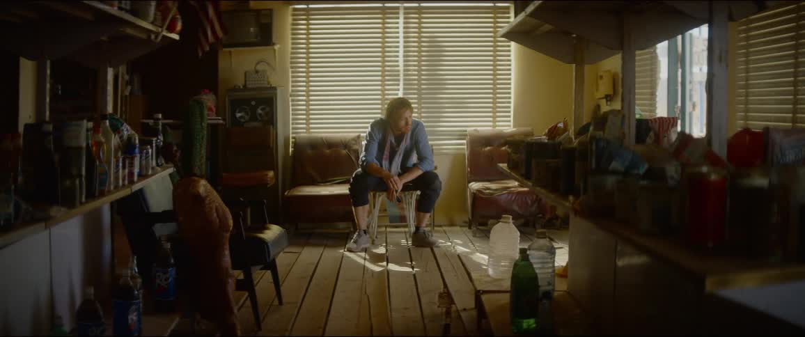 Man with a blue shirt and scarf sits in a chair and rests his elbows on his knees in a room
