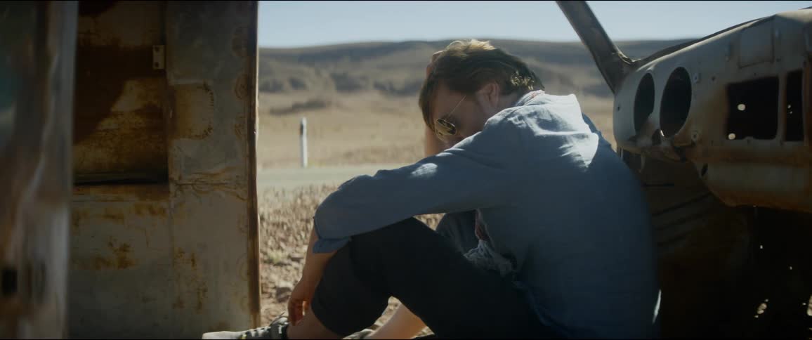 A man sits against a rusted and abandoned car in the desert