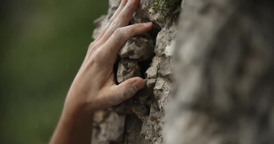 A hand reaches into frame attempting to grab the surface of a rock face but the rock crumbles beneath its grip and the hand falls out of frame