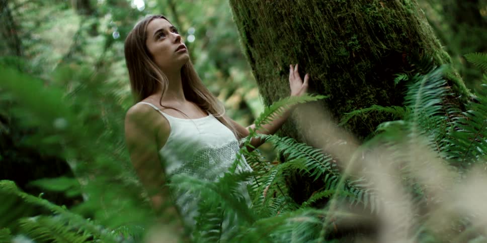 A teenager poses in a forest with her hand resting on a large tree trunk