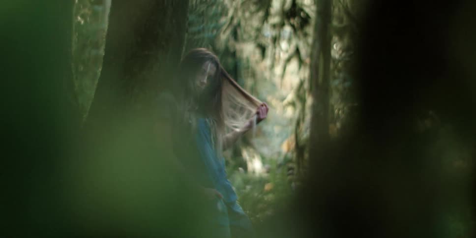 Woman leans against tree in forest wearing overalls and playing with hair