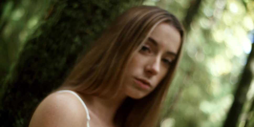 Woman with brown hair and a white dress leans against a tree in the forest