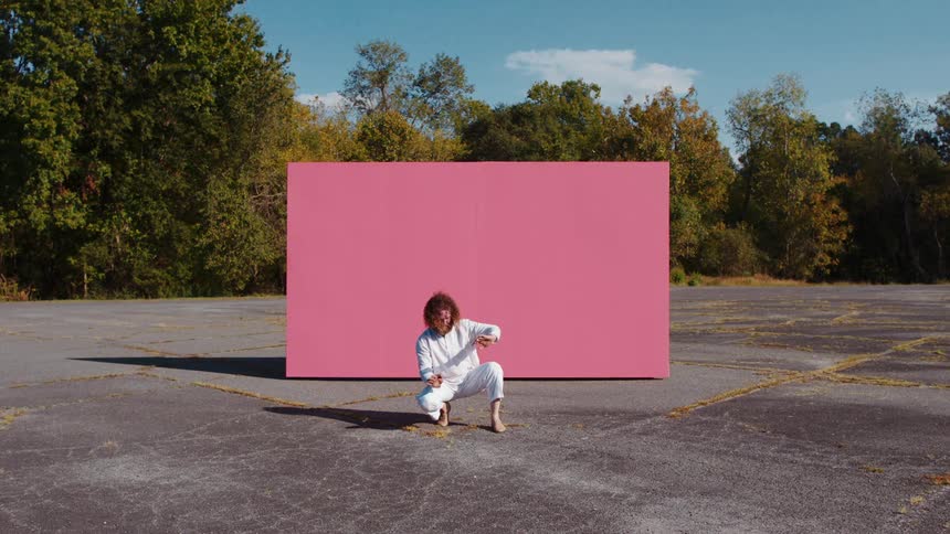A man kneels in front of a large pink box in a parking lot and raises his arms to block his eyes from the sun