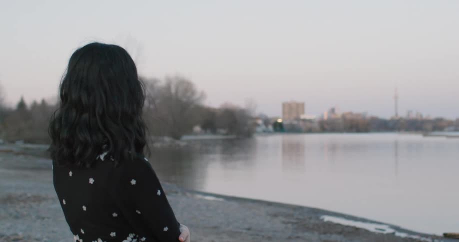 Woman in a long dress stands on the shoreline of a lake at sunset