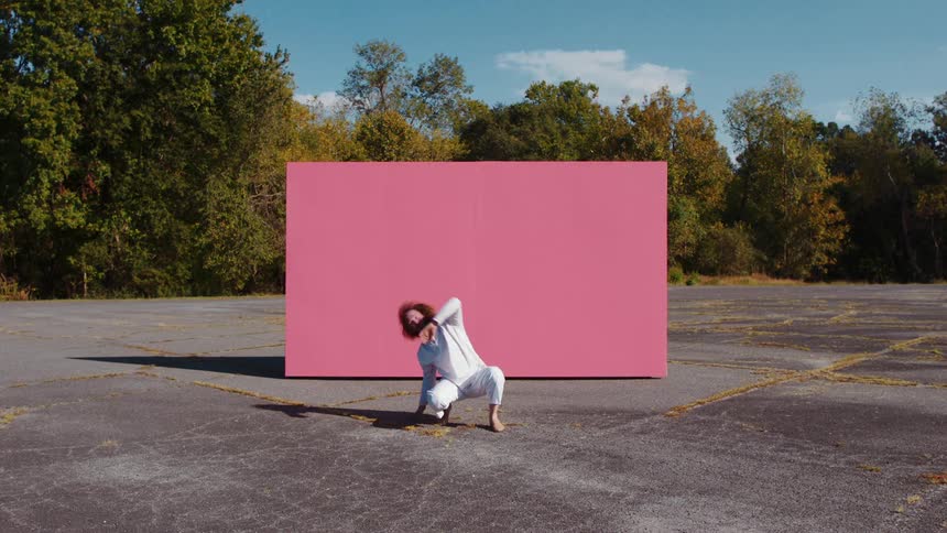 A white man dressed all in white performs an odd kneeling dance in front of a pink back drop