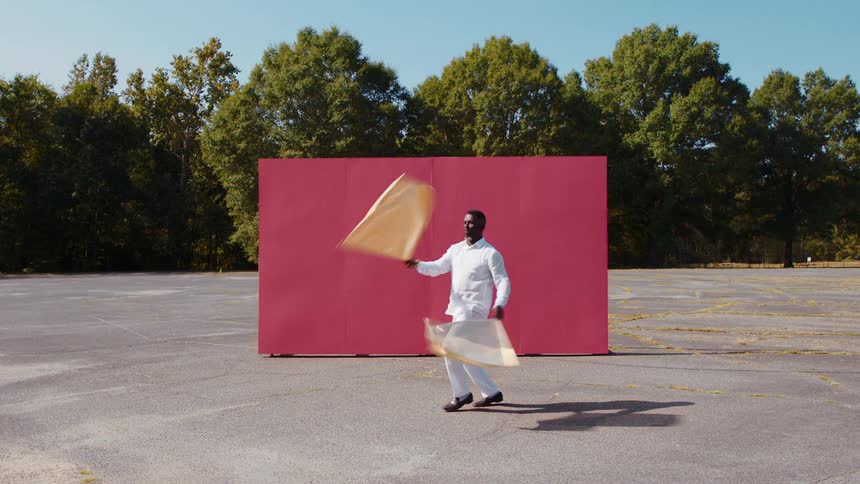 A black man waves flags and dances in front of a back drop in a parking lot