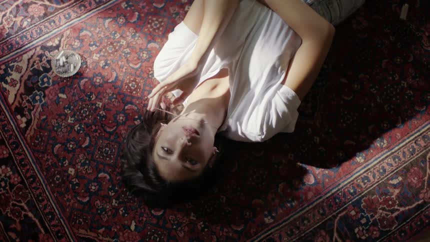 Young woman laying down on a rug on the floor, playing with her necklace, looking into camera