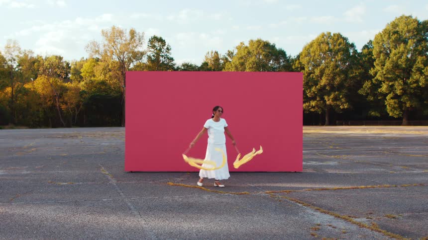Woman in a white dress dances in front of a red backdrop in an empty parking lot near a tree line at sunset