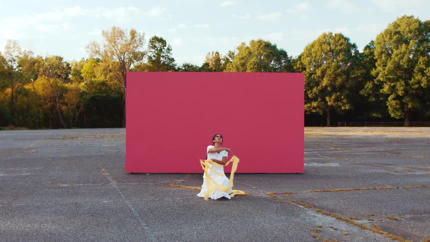 Woman in a white dress dances in front of a pink backdrop in a parking lot near a tree line