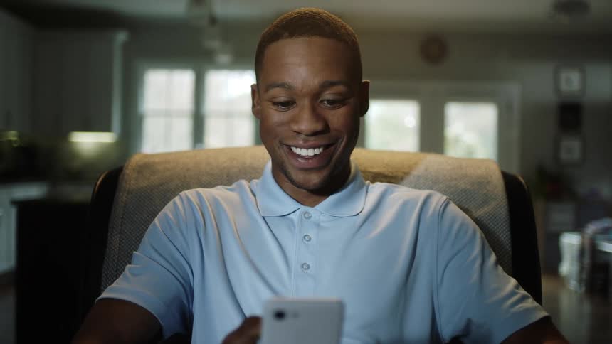 Happy black man with a blue polo shirt and sitting in an armchair uses a smartphone and bites his lips