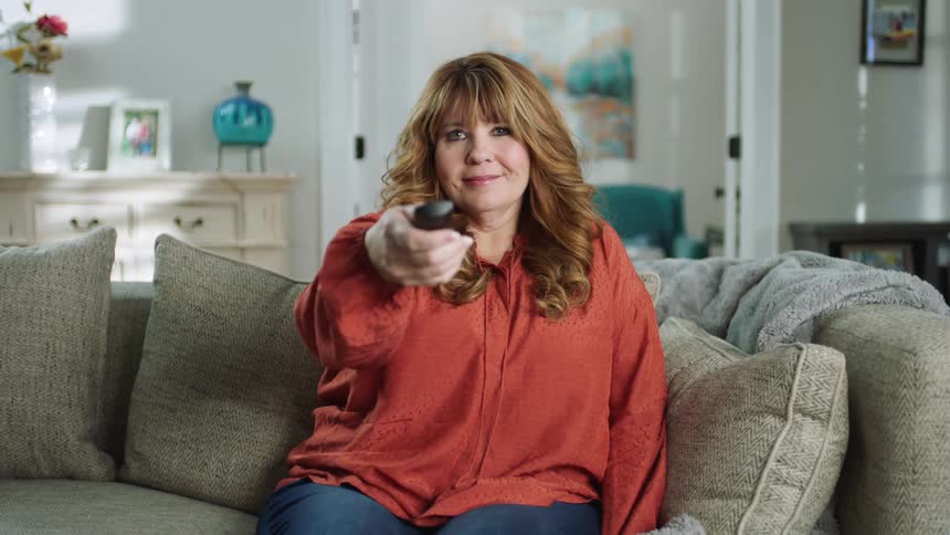 A woman sits down on her couch and turns on her tv remote control