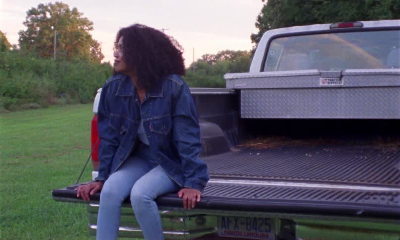 A woman sits in the bed of a pickup truck wearing jeans and a denim jacket