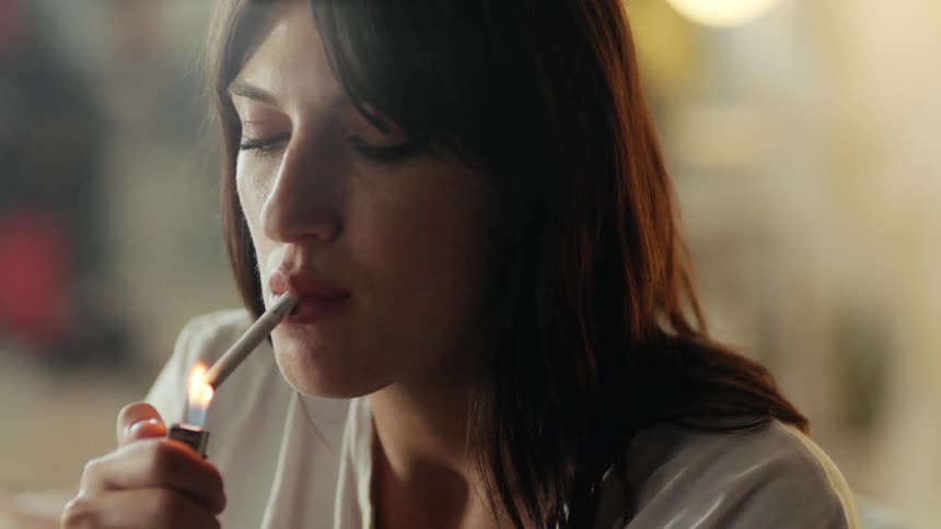 Woman with brown hair and a white shirt lights a cigarette inside a loft.