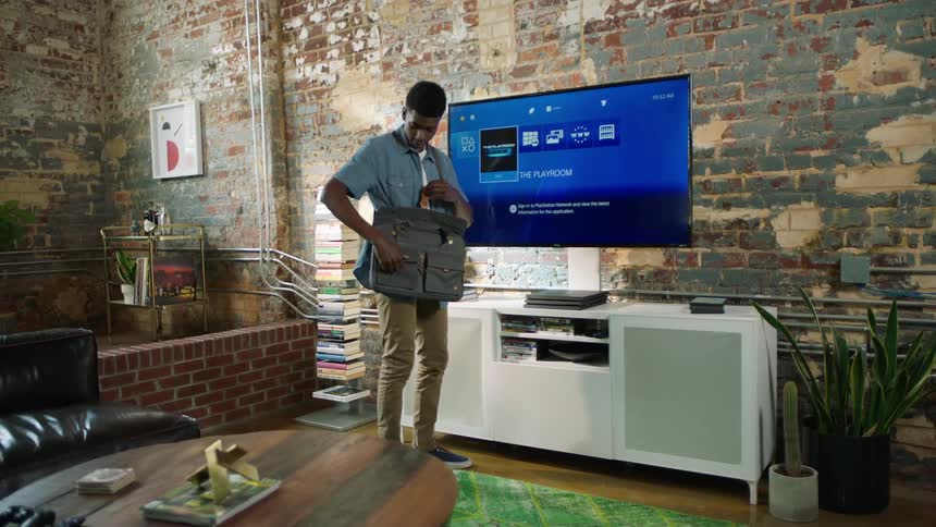 A young man stands in front of his television and looks through his messenger bag